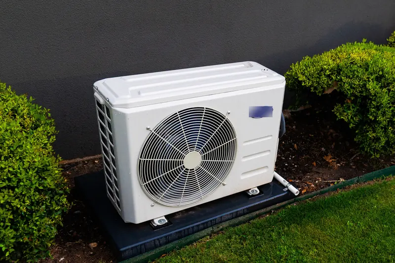 White heat pump outdoor unit on a black pad between clipped shrubs