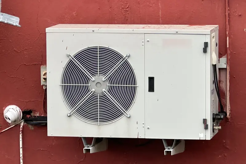 Grey air-conditioning outdoor unit on wall brackets against a red rendered house wall