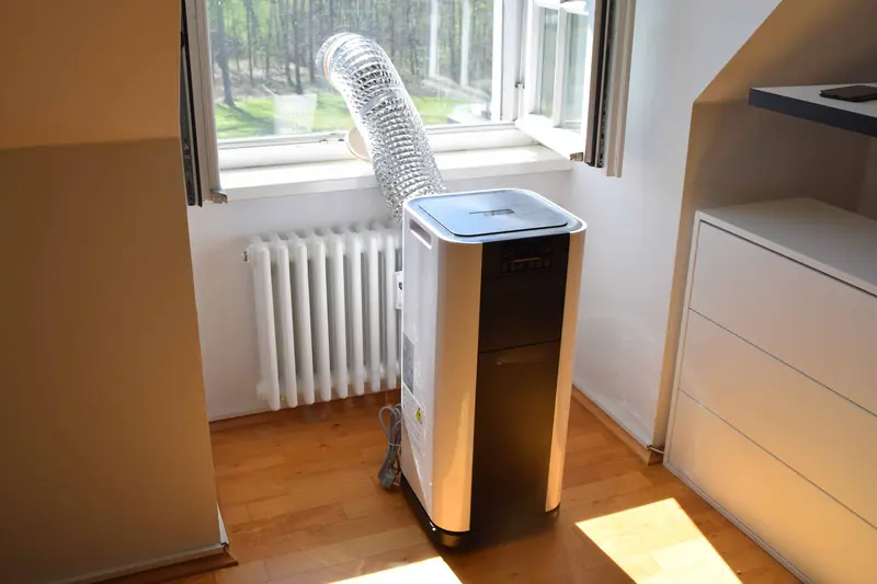 Portable air conditioner on a wooden floor with its silver exhaust hose running out of an open window